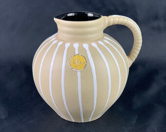 Può includere: Una brocca in ceramica beige con corpo rotondo e manico. La brocca è decorata con strisce bianche verticali e ha un interno nero. Una piccola etichetta gialla con testo è sul davanti.