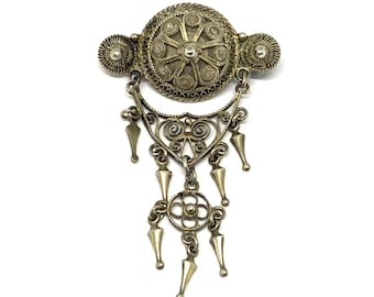 May include: A silver filigree brooch with a round central design and a dangling pendant with multiple points.