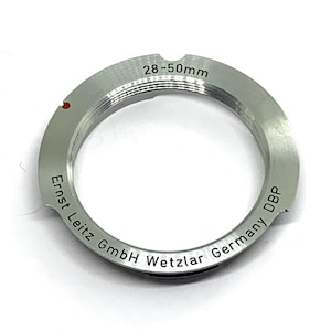 Op de afbeelding: Zilverkleurige camera lens adapterring met de tekst "28-50mm" en "Ernst Leitz GmbH Wetzlar Germany DBP" gegraveerd. De adapter heeft een rode stip en is ontworpen voor camera-apparatuur.