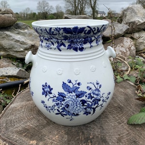 May include: A white ceramic planter with blue floral designs. The planter has two handles and a rounded body.