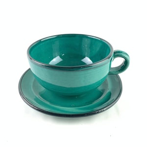May include: A green ceramic teacup and saucer set. The cup has a rounded shape and a handle. The saucer is round and has a slightly raised edge.