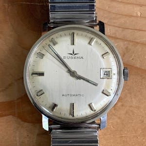 May include: Vintage DUGENA automatic wristwatch with a silver-tone metal band. The watch face is white with black numerals and hands, and a date window at the 3 o'clock position. The watch has the word "DUGENA" and "AUTOMATIC" printed on the face.
