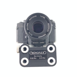 May include: A black camera with a silver lens and a black NOVOFLEX adapter plate. The lens is labeled "NOVOFLEX AUTO GERMANY 1:4/105 NOFLEXAR BELLOWS". The adapter plate is labeled "NOVOFLEX Germany D.B.Pat. U.S.Pat."