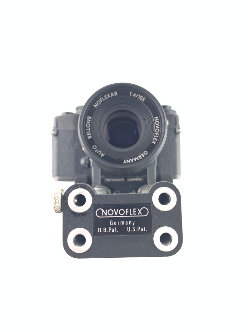May include: A black camera with a silver lens and a black NOVOFLEX adapter plate. The lens is labeled "NOVOFLEX AUTO GERMANY 1:4/105 NOFLEXAR BELLOWS". The adapter plate is labeled "NOVOFLEX Germany D.B.Pat. U.S.Pat."