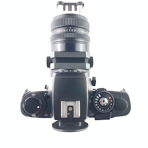 May include: A black 35mm film camera with a black lens attached. The camera has a black leather grip and a silver hot shoe. The lens has a black metal body and a silver ring with numbers on it.