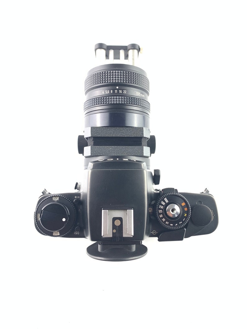 May include: A black 35mm film camera with a black lens attached. The camera has a black leather grip and a silver hot shoe. The lens has a black metal body and a silver ring with numbers on it.