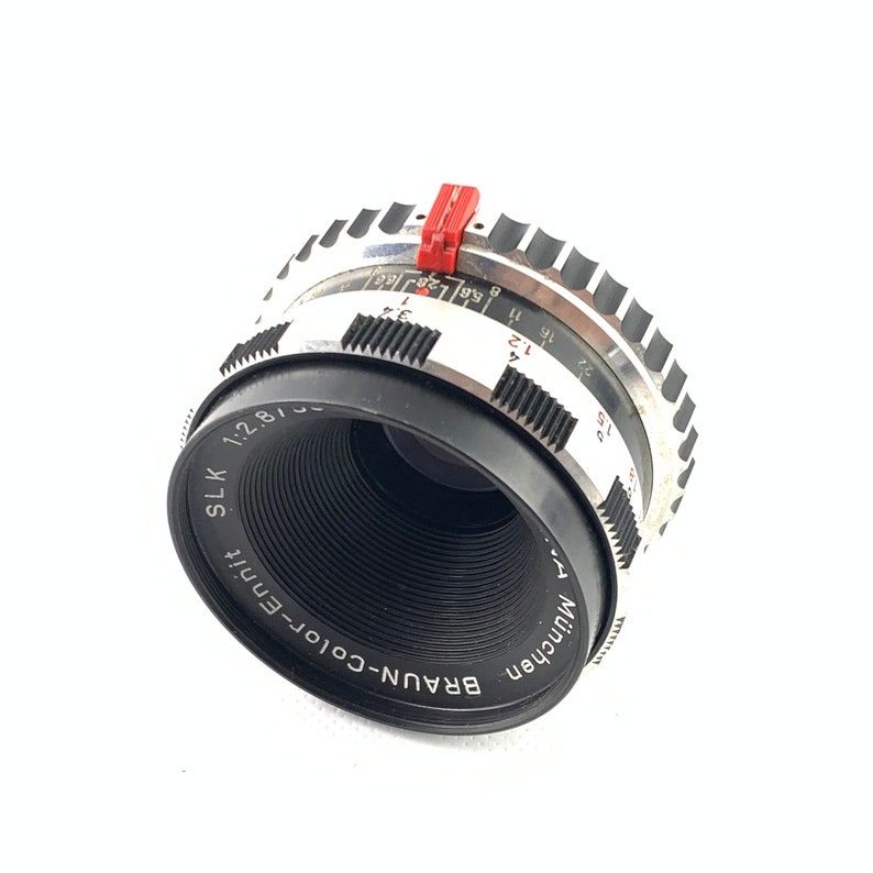 K&ouml;nnte beinhalten: Ein silbernes und schwarzes Braun SLK 1:2.8/50mm Kameraobjektiv mit einem roten Blendenring. Das Objektiv hat einen schwarzen Lauf mit wei&szlig;em Text, der "BRAUN-Color-Ennit M&uuml;nchen" und "SLK 1:2.8/50mm" lautet.