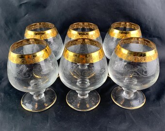 Puede incluir: Seis copas de brandy transparentes con bordes dorados y grabados decorativos. Las copas tienen una forma de cuenco redondeada y se asientan sobre un tallo corto. El borde dorado presenta un patrón ornamental repetido.