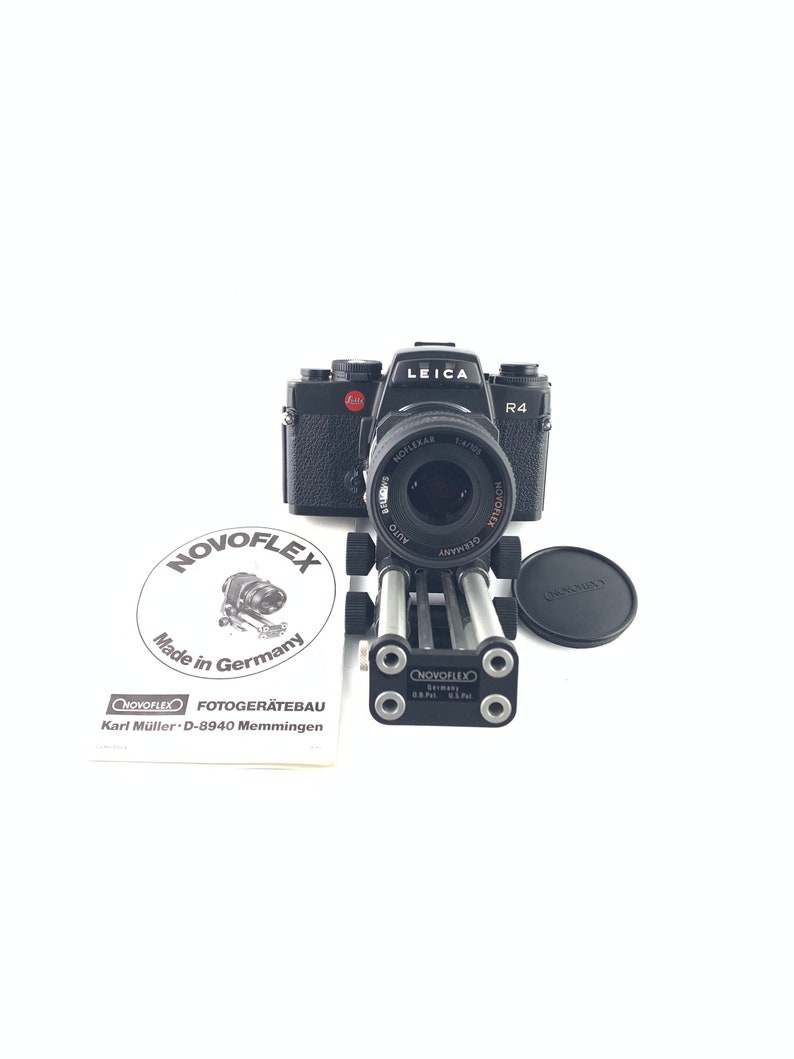 May include: A black Leica R4 camera with a silver lens attached to a black camera tripod. The camera is on a white background. The tripod is made by NOVOFLEX and is made in Germany. The camera has a red dot on the top and the lens has the text "HOFLEXAR 1:4/105 AUTO BELOVE" printed on it.