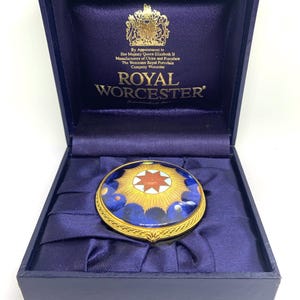 Puede incluir: Un joyero redondo de porcelana Royal Worcester con borde de metal dorado y un diseño de rayos de sol en azul, amarillo y rojo en la tapa. La caja se presenta en un estuche azul oscuro con texto dorado y un escudo real impreso en el interior.
