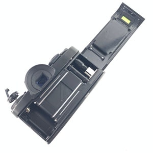 May include: A black film camera with the back open, revealing the film compartment and a small green light. The camera is lying on a white surface.