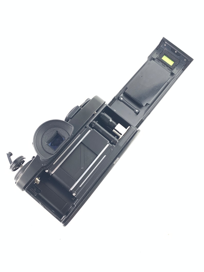 May include: A black film camera with the back open, revealing the film compartment and a small green light. The camera is lying on a white surface.