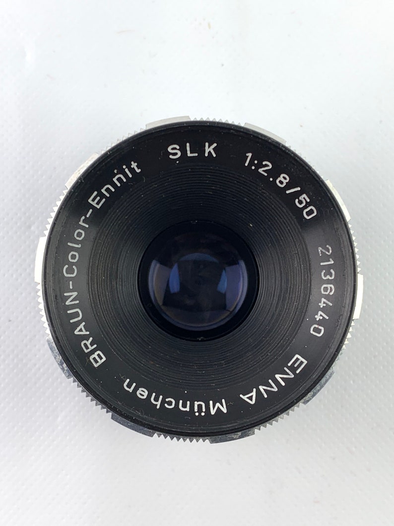 K&ouml;nnte beinhalten: Ein schwarzes und silbernes Kameraobjektiv mit dem Text "ENNA 0 171362", "BRAUN-COLOR-Ennit", "MUNchen", "SLK 1:2.8/50" um den Rand gedruckt.