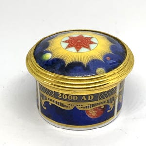 Puede incluir: Una pequeña caja decorativa redonda con borde y tapa dorados. La tapa presenta un diseño celestial con un sol dorado y una estrella roja. La caja es azul oscuro con el texto "2000 AD" y detalles dorados.