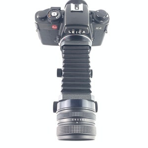 May include: A black Leica R4 camera with a bellows extension tube and a black lens. The camera has a red "Leica" logo and a black "R4" label.