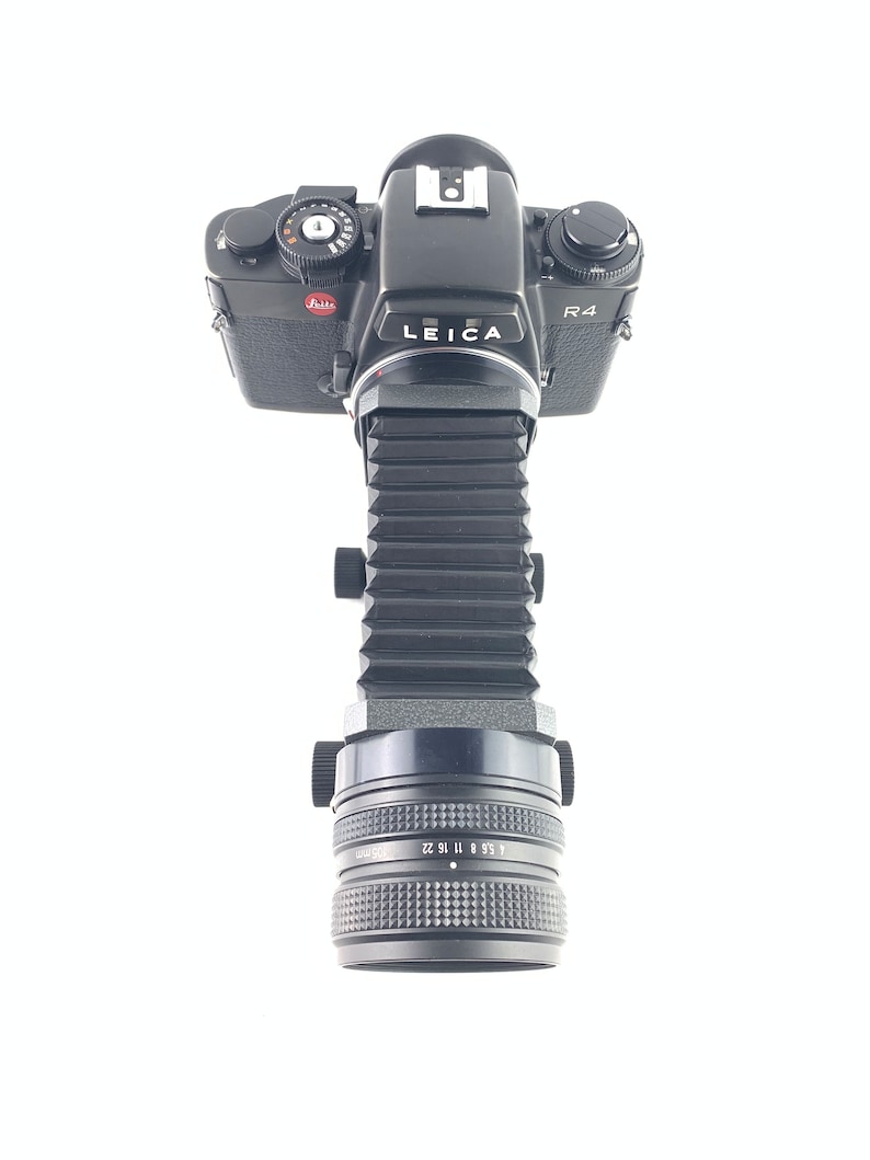May include: A black Leica R4 camera with a bellows extension tube and a black lens. The camera has a red "Leica" logo and a black "R4" label.