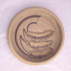 May include: A beige ceramic bowl with a brown and beige design of three stylized leaves. The bowl has a slightly raised rim.