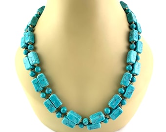 Op de afbeelding: Een turquoise ketting op een mannequin. De ketting heeft rechthoekige en ronde turquoise kralen, verbonden door goudkleurige metalen accenten. De ketting is dubbelstrengs en heeft een vintage uitstraling.