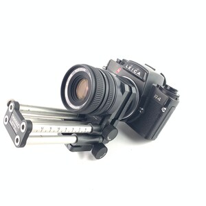 May include: A black Leica R4 camera with a black lens attached to a silver sliding rail system. The camera has a red "LEICA" logo and "R4" text on the body.