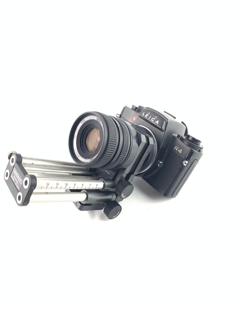 May include: A black Leica R4 camera with a black lens attached to a silver sliding rail system. The camera has a red "LEICA" logo and "R4" text on the body.