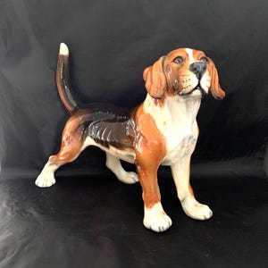 May include: A glossy ceramic beagle figurine standing with its tail held high. The dog features a tri-color coat with patches of brown, black, and white, with a white chest and paws, and a black saddle pattern across its back.