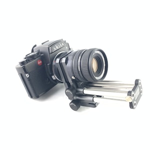 May include: A black Leica camera with a lens attached to a tripod. The camera has a red "Leica" logo on the top.