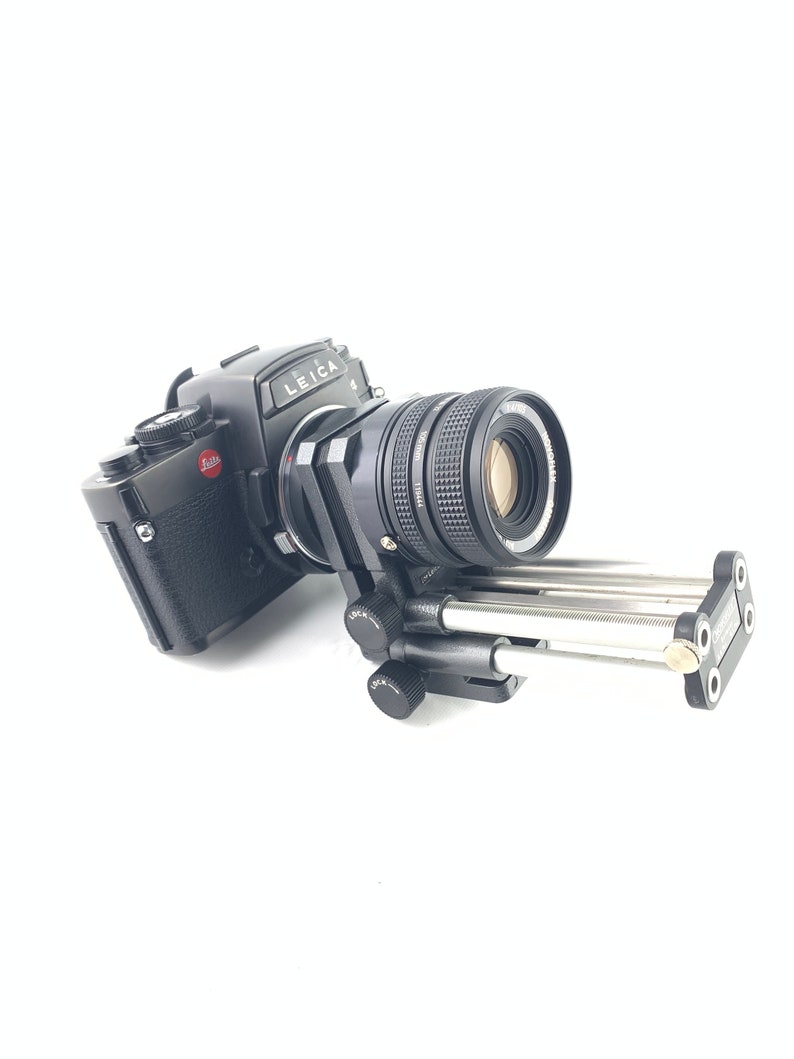 May include: A black Leica camera with a lens attached to a tripod. The camera has a red "Leica" logo on the top.