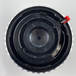 K&ouml;nnte beinhalten: Nahaufnahme eines schwarzen Kameraobjektivs mit einem silbernen Ring und einem roten Hebel. Das Objektiv hat eine kreisf&ouml;rmige Blende mit 7 Bl&auml;ttern.