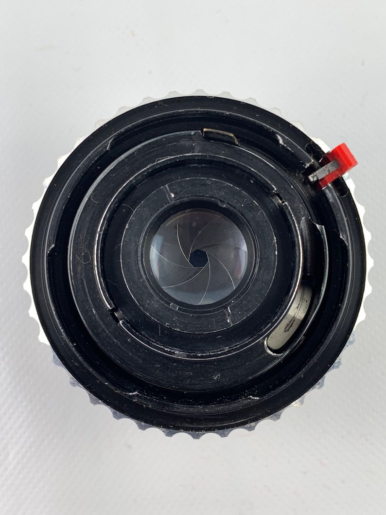K&ouml;nnte beinhalten: Nahaufnahme eines schwarzen Kameraobjektivs mit einem silbernen Ring und einem roten Hebel. Das Objektiv hat eine kreisf&ouml;rmige Blende mit 7 Bl&auml;ttern.