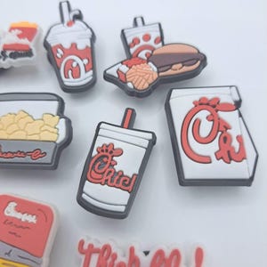 May include: A collection of small, colorful rubber shoe charms featuring fast food themes. The set includes miniature representations of a drink cup, a burger with fries, a popcorn container, and a white charm with the red text Thick-fil-A.