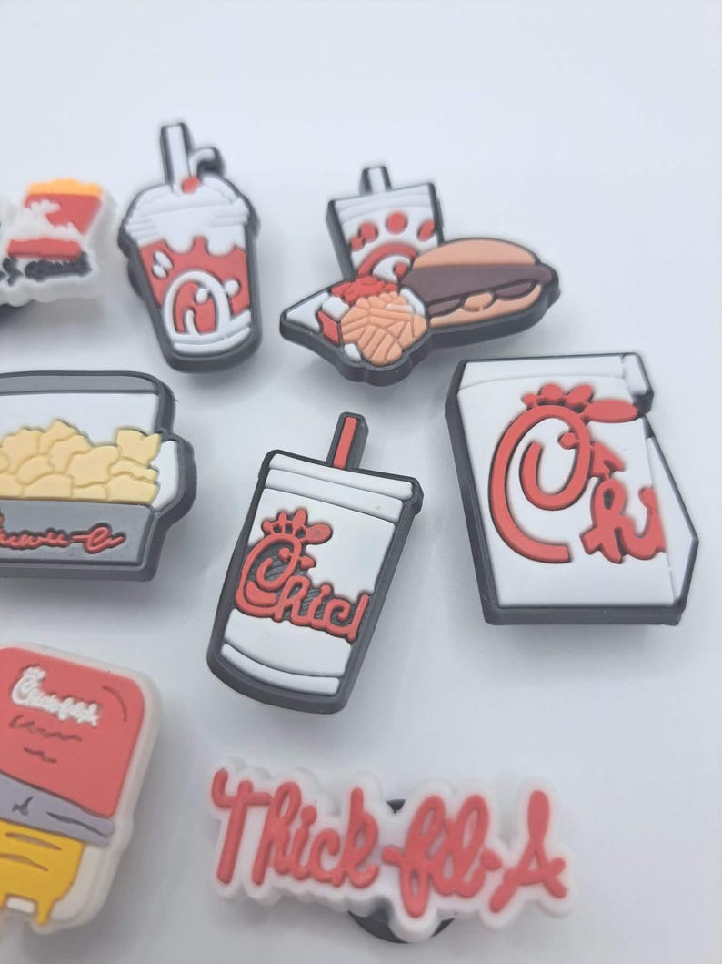 May include: A collection of small, colorful rubber shoe charms featuring fast food themes. The set includes miniature representations of a drink cup, a burger with fries, a popcorn container, and a white charm with the red text Thick-fil-A.