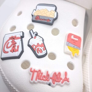 May include: Five decorative shoe charms attached to a white foam clog. The charms feature a box of nuggets, a drink cup, a fry container, a sauce packet, and a white charm with red text that reads Thick-fil-A in a stylized script font.