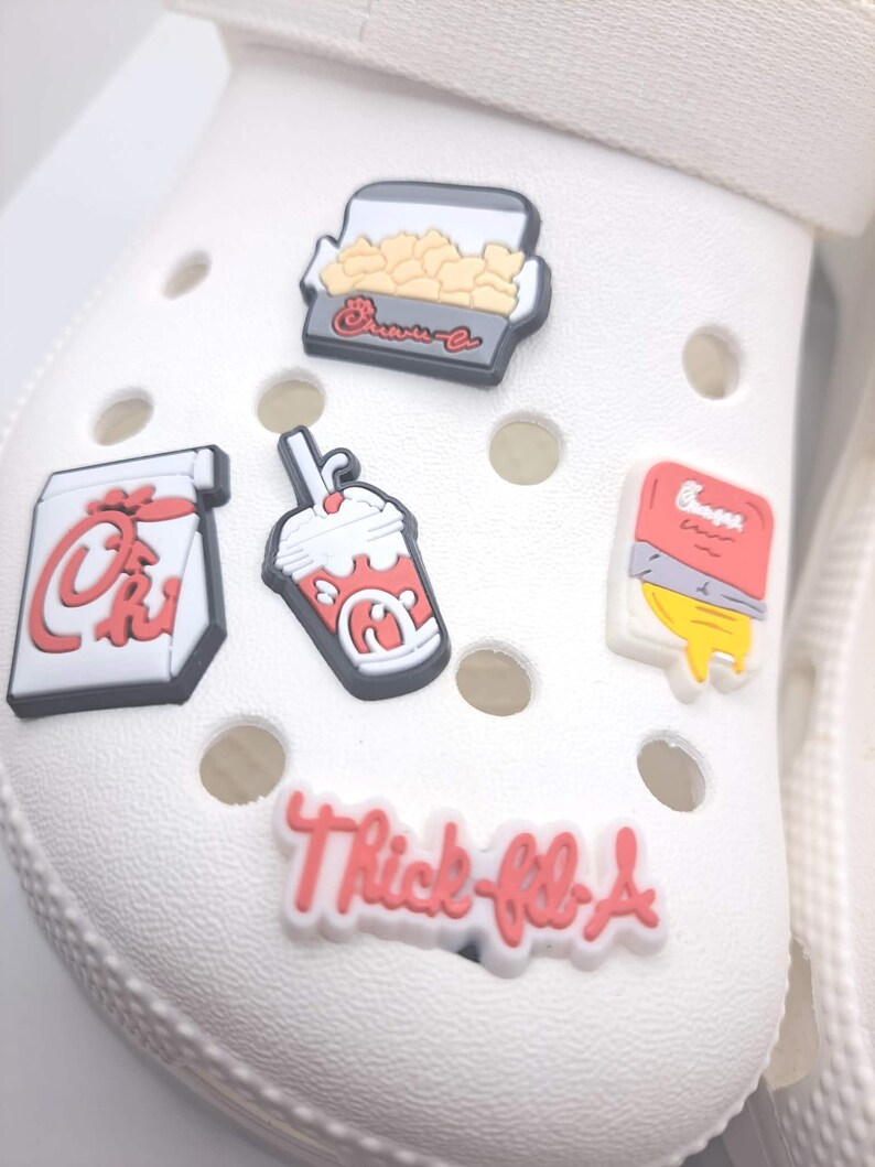 May include: Five decorative shoe charms attached to a white foam clog. The charms feature a box of nuggets, a drink cup, a fry container, a sauce packet, and a white charm with red text that reads Thick-fil-A in a stylized script font.