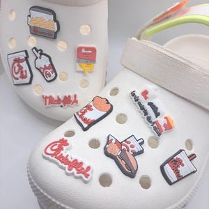 May include: A pair of white foam clogs decorated with various colorful rubber charms featuring fast food icons, including drink cups, sandwich boxes, and fries, along with the Chick-fil-A logo and text in red, white, and black colors.