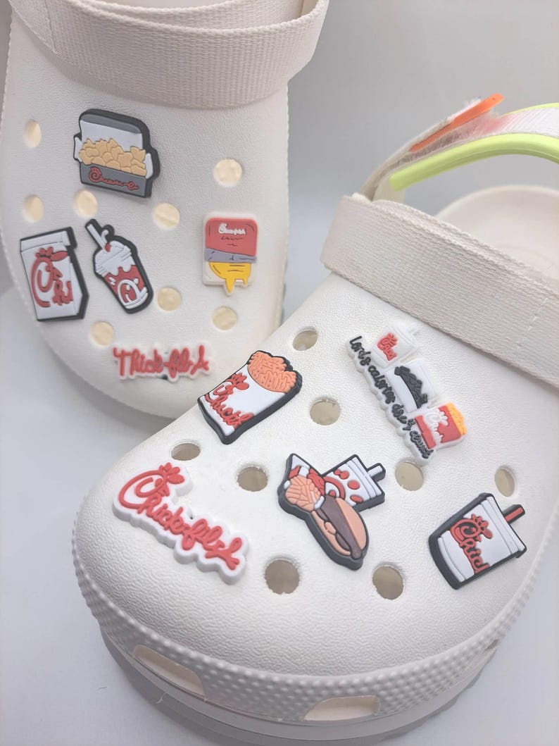 May include: A pair of white foam clogs decorated with various colorful rubber charms featuring fast food icons, including drink cups, sandwich boxes, and fries, along with the Chick-fil-A logo and text in red, white, and black colors.
