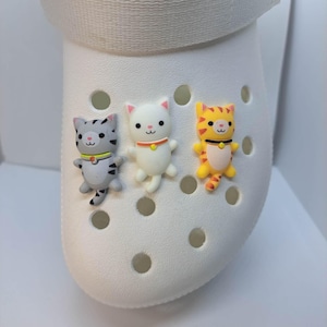 May include: White shoe with three cat-shaped charms attached. The charms are gray, white, and orange, each with a collar. The shoe has multiple holes and a white strap.
