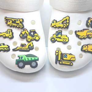 May include: White clogs decorated with yellow and black construction vehicle charms, including excavators, dump trucks, and a crane. A green dump truck charm is also present. These charms add a playful touch to the footwear.