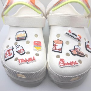 May include: A pair of white foam clogs featuring adjustable straps with neon yellow accents. The shoes are decorated with various colorful rubber charms depicting fast food items, including containers of fries, a sandwich, and Chick-fil-A logos.