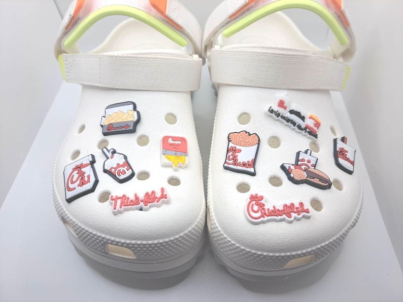 May include: A pair of white foam clogs featuring adjustable straps with neon yellow accents. The shoes are decorated with various colorful rubber charms depicting fast food items, including containers of fries, a sandwich, and Chick-fil-A logos.