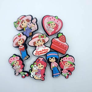 May include: A collection of nine colorful rubber shoe charms featuring Strawberry Shortcake characters, a strawberry-topped cake, and a heart-shaped frame. The designs include various figures with pink hats, red hair, and pets, with one charm displaying the text Strawberry Shortcake.