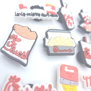 May include: A collection of rubber shoe charms featuring various fast food icons, including a fry container, a drink cup, and a nugget box, all decorated with red and white branding and the Chick-fil-A logo, with one charm displaying the text Lord's calories.