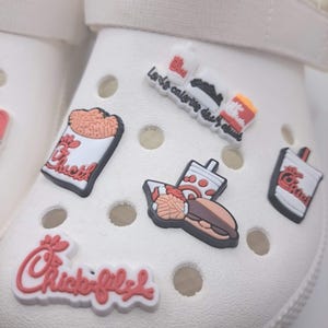 May include: A white foam clog shoe decorated with multiple Chick-fil-A themed rubber charms. The charms feature the brand logo, a sandwich, a drink, and a container of waffle fries. The shoe has a bright neon yellow heel strap and circular vents.