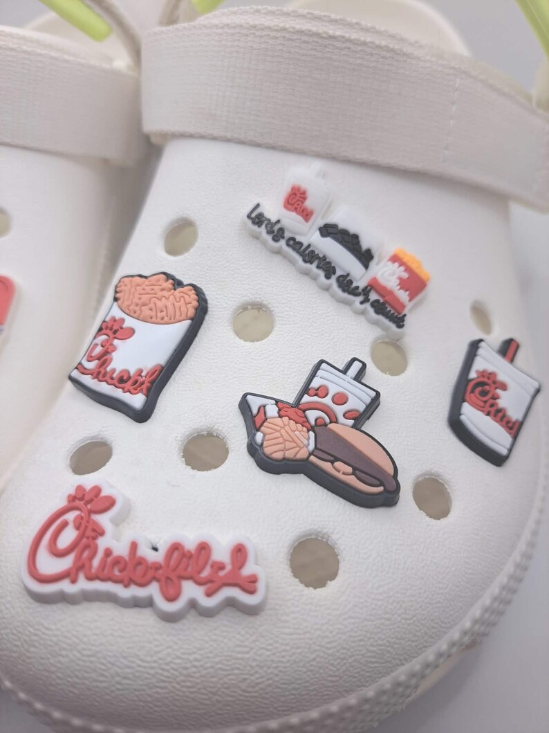 May include: A white foam clog shoe decorated with multiple Chick-fil-A themed rubber charms. The charms feature the brand logo, a sandwich, a drink, and a container of waffle fries. The shoe has a bright neon yellow heel strap and circular vents.