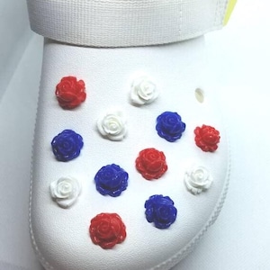 May include: White sandal with a white strap and orange accents. The sandal is decorated with red, white, and blue rose-shaped charms. The sandal is a casual footwear item.