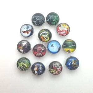 May include: A collection of twelve circular metal snap buttons featuring various horror-themed graphic prints. Each button displays distinct imagery including skulls, monsters, movie-inspired artwork, and text such as Night of the Living Dead and Screaming Skull, all set under a clear domed finish.