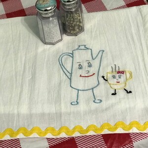 May include: A white tea towel with yellow scalloped edging features an embroidered design of a teapot and a teacup with faces. The teapot and teacup are both wearing a smile and have eyes. The teacup has a red bow on its lid. Two clear glass salt and pepper shakers are on top of the towel.