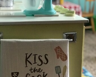 May include: A white kitchen towel with the text "Kiss the Cook" in brown lettering. The towel has a brown and white illustration of a spatula, a brown and white illustration of a pot, and a brown and white illustration of a mitten.
