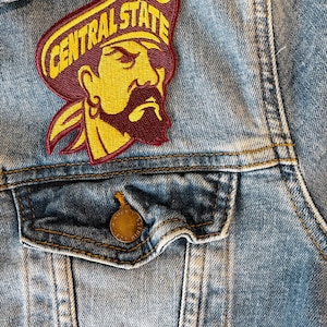 May include: A maroon and gold embroidered patch with the text "CENTRAL STATE" and a cartoon pirate with an eyepatch. The patch is sewn onto a blue denim jacket.