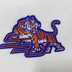 May include: Embroidered patch featuring a tiger in orange and white stripes, set against a blue background with the letters "SJ". The patch has a blue border and is likely for apparel or accessories.