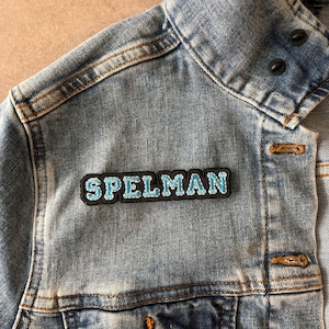May include: A blue denim jacket with a black and blue embroidered patch that says "SPELMAN" on the left shoulder.
